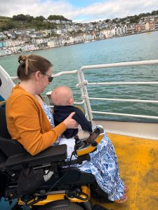 Dr Anna Severwright sat on a ferry, in a motorised wheelchair with her son Theo sitting on her lap and they are both looking out at the view of a harbour.