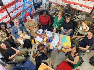 A group of people sat around a table in a working area with bookshelves and files. All the people are looking up and smiling at the camera.